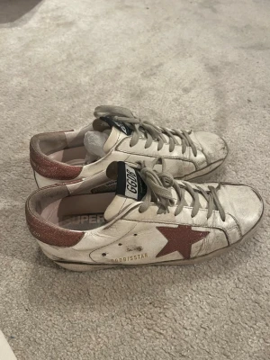 Golden Goose vita & rosa sneakers  - Snygga vita sneakers från Golden Goose med rosa stjärna på sidan och rosa glitterdetalj på hälen. Skorna har vit/gråa skosnören, svart GGDB-märke på plösen och är tillverkade i läder. Perfekta för en cool och avslappnad stil. Köpta på Vestiaire där dem har gjort en äkthetskontroll💕 Jag har fler bilder, det är bara att fråga om jag ska skicka dem!