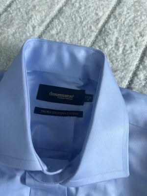 Ljusblå skjorta från Dressmann - Klassisk ljusblå skjorta från Dressmann i tvålagers egyptisk bomull. Skjortan har knappar framtill och en stilren krage, perfekt för en clean och snygg look. Passar dig som gillar enkel och tidlös stil.