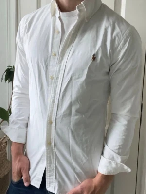 Vit skjorta från Ralph Lauren - Klassisk vit skjorta från Ralph Lauren med button-down krage och broderad logga på bröstet. Skjortan har lång ärm, knappar framtill och är tillverkad i bomull. Perfekt för en clean och stilren look.