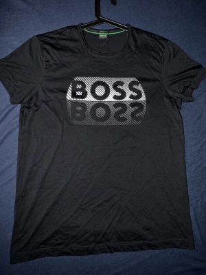 Svart BOSS t-shirt med logga - Svart t-shirt från BOSS med stor, glansig logga framtill i silverton. Klassisk rund hals och regular fit. Perfekt för dig som gillar stilrena och trendiga plagg med tydlig branding.