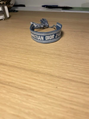 Blått Christian Dior J'Adior armband - Snyggt blått tygarmband från Christian Dior med texten 'J'Adior' broderad i vitt. Armbandet har justerbar knytning med tofsar i ändarna och en cool, ungdomlig vibe. Perfekt accessoar för att lyfta din outfit med en touch av lyx.