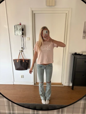 Ljusblå slitna bootcut jeans - Säljer ett par ljusblå jeans med bootcut passform och slitningar vid knäna. Jeansen har klassisk femficksdesign och är tillverkade i denim med lite stretch för extra komfort. Perfekta för en avslappnad och trendig look.