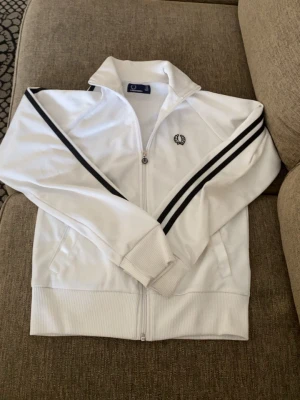 Vit Fred Perry ziptröja med svarta ränder - Snygg vit ziptröja från Fred Perry med svarta ränder längs ärmarna och broderad logga på bröstet. Tröjan har hög krage, ribbade muddar och dragkedja framtill. Perfekt för en sportig och stilren look.