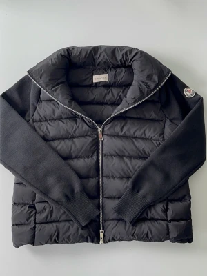 Svart pufferjacka från Moncler - Svart pufferjacka från Moncler med quiltad front och rygg, stickade ärmar och hög krage. Jackan har dragkedja framtill och Moncler-logga på ena ärmen. Perfekt för kalla vinterdagar och har en modern, stilren look.