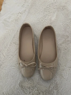 Beige ballerinaskor med rosett - Snygga beige ballerinaskor i glansigt material med rund tå och söt rosettdetalj framtill. Skorna har en klassisk och enkel design som passar till många olika outfits. Perfekta för dig som gillar stilrena och feminina skor.