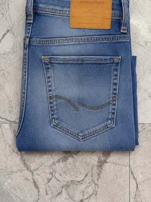Blå slitna jeans från Jack & Jones - Säljer ett par blå jeans från Jack & Jones med snygga slitningar på låren och knäna. Jeansen har klassisk femficksdesign, ljus tvätt och raka ben. Materialet är denim och bakfickan har dekorativ söm. Perfekt för en avslappnad look.