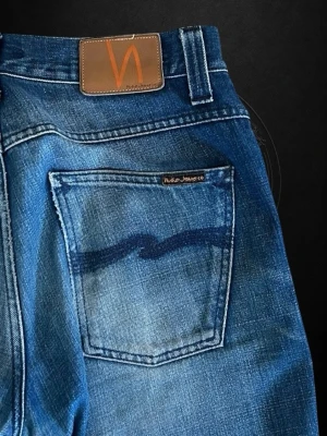 Blå salvedge jeans från Nudie Jeans - Snygga blå jeans från Nudie Jeans Co med klassisk femficksdesign och ikonisk våg på bakfickan. Jeansen har en rak passform och är tillverkade i denim av bomull. Perfekta för en avslappnad och trendig look. 