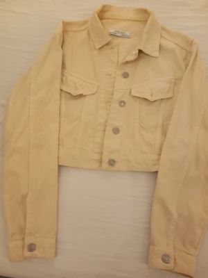 Beige cropped jeansjacka - Snygg beige cropped jeansjacka från dilvin jeans Woman med klassisk krage, silverfärgade knappar och två bröstfickor med lock. Jackan har raka ärmar och är tillverkad i ett mjukt denimtyg. Perfekt för en trendig och avslappnad look.