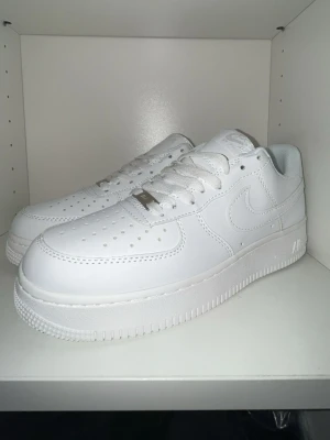 Nike Air Force 1 vita sneakers - Klassiska Nike Air Force 1 sneakers i helvitt läder med perforerad tå, vit sula och ikonisk Swoosh på sidan. Skorna har lågt skaft och vita skosnören. Perfekta för en clean och stilren look.