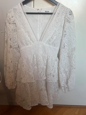 Vit spetsklänning från Showpo - Superfin vit kort klänning från Showpo med v-ringning och långa ärmar. Klänningen har vackra spetsdetaljer och volanger, vilket ger en romantisk och trendig look. Perfekt för sommarens alla tillfällen!