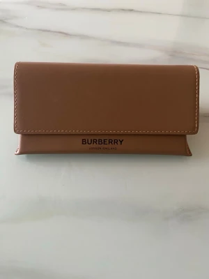 Burberry solglasögonfodral beige - Stilrent solglasögonfodral från Burberry i beige skinn med klassisk logga framtill. Medföljer matchande tygpåse och putsduk, båda i samma beige nyans och med Burberry-tryck. Perfekt för att förvara och skydda dina solglasögon.