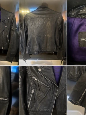Svart skinnjacka Rockandblue - Snygg svart skinnjacka från Rockandblue med klassisk bikerstil, silvriga dragkedjor och detaljer. Fodrad med lila tyg på insidan. Jackan har flera fickor med dragkedja och coola axelpartier.