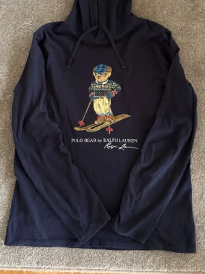 Mörkblå hoodie Polo Bear Ralph Lauren - Mörkblå hoodie från Ralph Lauren med Polo Bear-tryck där björnen åker skidor. Långärmad modell med huva och dragsko. Tillverkad i mjukt bomullsmaterial, perfekt för en avslappnad och cool stil.