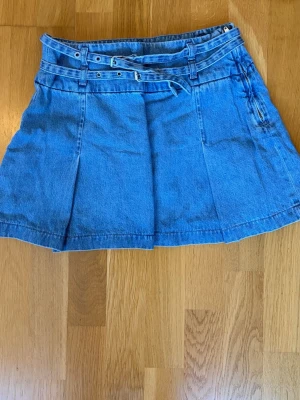 Blå jeanskjol med bälte från Zara - Snygg blå jeanskjol från Zara i kort modell med dubbla bälten och hällor i midjan. Kjolen har en klassisk denimlook och är perfekt för dig som gillar en trendig och avslappnad stil. Katt finns i hemmet!