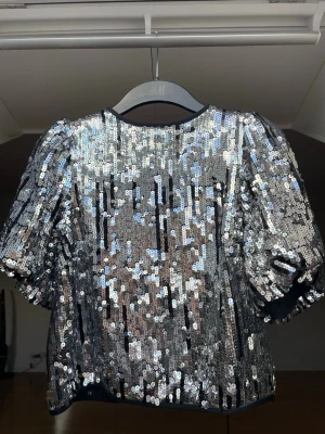 Glittrig topp  - Snygg topp från H&M täckt med silvriga paljetter. Den har puffiga korta ärmar och rund halsringning. Koftan har knäppning baktill och är fodrad på insidan.