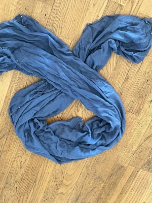Blå tunn sjal i bomull - En snygg blå sjal i tunt, mjukt tyg som ger en avslappnad och trendig look. Perfekt att vira runt halsen eller använda som accessoar till jackan. Materialet känns lätt och luftigt, och färgen är en dämpad blå som passar till det mesta.
