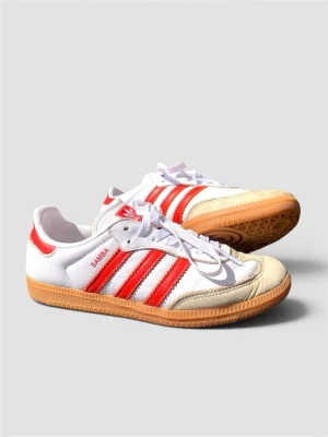 Adidas Samba vita/röda sneakers - Klassiska Adidas Samba sneakers i vitt läder med röda detaljer och tre röda ränder på sidorna. Skorna har vit ovandel, beige mockadetaljer vid tån och gummisula. Perfekt för dig som gillar retrostil och streetwear.