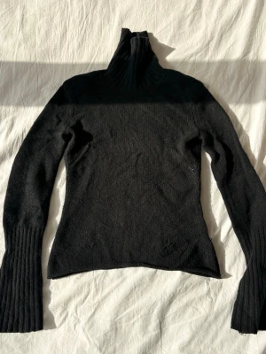 Svart stickad polotröja - Svart stickad polotröja med långa ärmar och breda ribbade muddar. Tröjan har en hög polokrage och är figurnära i modellen. Perfekt för lager-på-lager och enkel att matcha med olika outfits.
