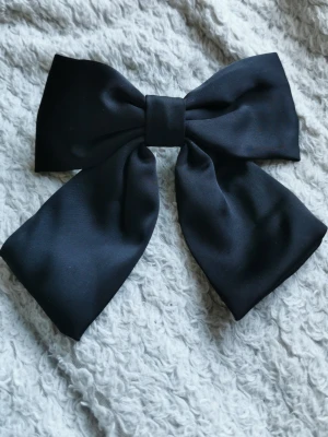 Stor svart hårrosett i satin - Snygg och stor svart hårrosett i glansigt satinmaterial. Rosetten har en klassisk form och sitter fast på ett metallspänne som gör den enkel att fästa i håret. Perfekt för att ge din frisyr en extra touch.