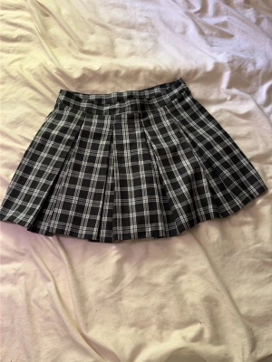 Svartvit rutig plisserad kortkjol - En svartvit rutig plisserad kortkjol med klassisk skoluniforms-vibe. Kjolen har bred linning och tydliga veck som ger extra volym. Perfekt för dig som gillar en trendig och ungdomlig stil.
