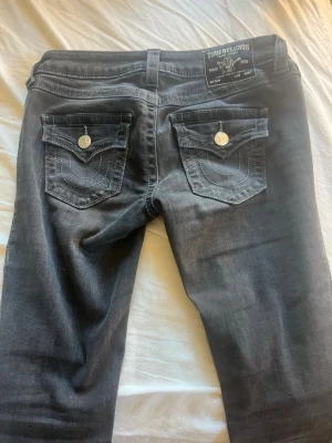 Gråa  jeans från True Religion - Snygga svarta jeans från True Religion med klassiska bakfickor och silverfärgade knappar. Jeansen har en tvättad look Perfekta för dig som gillar streetstyle och vill ha något som sticker ut lite extra. Använda 3-4 gånger och absolut ingenting fel med dem. Säljer då dem är för korta för mig. 
