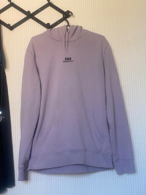 Ljuslila hoodie från Helly Hansen - Ljuslila hoodie från Helly Hansen med svart logga på bröstet och stor Helly Hansen-tryck på ryggen. Klassisk känguruficka framtill, huva med dragsko och ribbade muddar. Perfekt för en chill och avslappnad stil.
