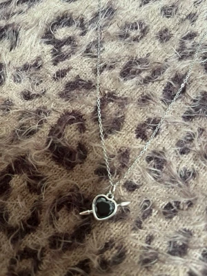 Halsband med svart hjärthänge silver - Snyggt halsband i silverfärgad metall med ett svart hjärthänge som har en pil genom sig. Kedjan är tunn och smidig, och hänget ger en cool och romantisk vibe. Perfekt accessoar för att lyfta din outfit med en unik detalj. Rostfri 