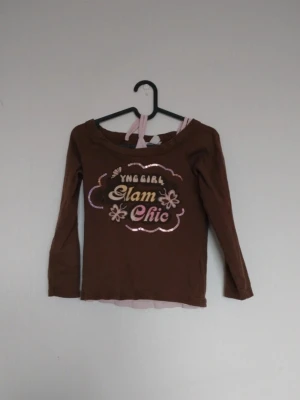 Brun långärmad topp med tryck - Brun långärmad topp med texten 'YNG GIRL Glam Chic' i glittrigt tryck framtill och blommiga detaljer. Rund halsringning och figurnära passform. Perfekt för dig som gillar en trendig och söt stil.