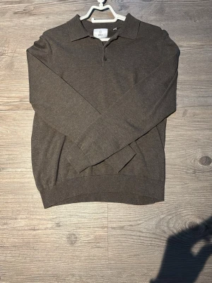 Brun pikétröja från Bläck - Snygg brun långärmad pikétröja från Bläck med klassisk krage och knäppning framtill. Tröjan är tillverkad i ett mjukt och tunt material som passar perfekt för lager-på-lager. Enkel och stilren design som funkar till många outfits.
