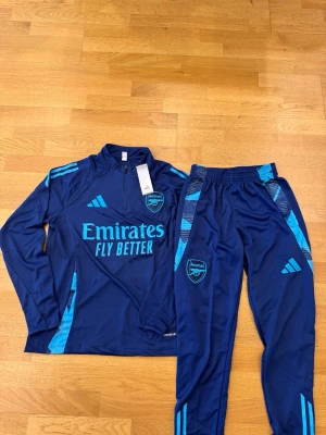 Arsenal träningsset i mörkblått - Snyggt Arsenal träningsset från Adidas i mörkblått med ljusblå detaljer. Setet består av en långärmad tröja med halv dragkedja och matchande byxor med resår i midjan. Båda plaggen har Arsenal-logga och Adidas-märke samt trycket 'Emirates Fly Better'. Perfekt för fotbollsträning.