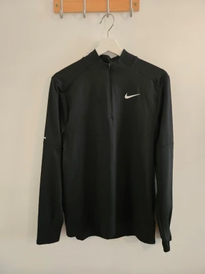 Svart långärmad löpartröja Nike - Svart långärmad löpartröja från Nike med halv dragkedja framtill och vit logga på bröstet. Tröjan har hög krage och är tillverkad i ett lätt och stretchigt funktionsmaterial som passar perfekt för träning och löpning.