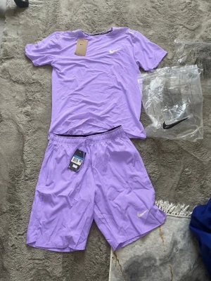 Lila träningsset från Nike - Snyggt lila träningsset från Nike med t-shirt och shorts. Båda plaggen har den klassiska Nike-loggan i vitt. Tillverkade i ett lätt och snabbtorkande material som passar perfekt för träning eller sportaktiviteter.