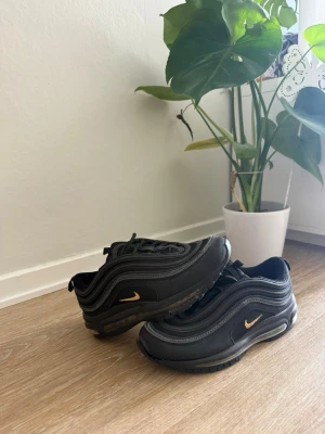 Svarta Nike Air Max 97 sneakers - Säljer ett par svarta Nike Air Max 97 sneakers med guldfärgade swoosh-detaljer. Skorna har den klassiska vågiga designen, synlig Air-sula och är tillverkade i en mix av läder och textil. Perfekta för dig som gillar streetwear och sportig stil.