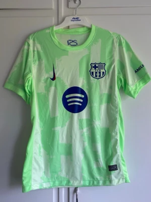Barcelona matchtröja ljusgrön Nike - Säljer en ljusgrön FC Barcelona matchtröja från Nike med klubbmärke och Spotify-logga på bröstet. Tröjan har korta ärmar, rund hals och UNHCR-tryck på ryggen. Tillverkad i lätt och ventilerande material, perfekt för fotboll eller träning.