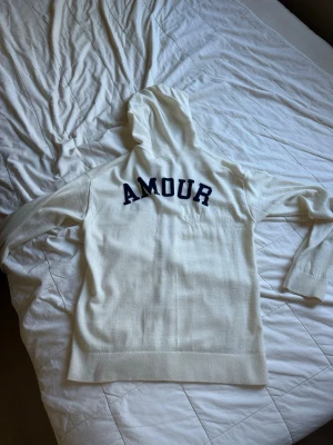 Zadig Voltaire amour hoodie - Säljer en vit hoodie med dragkedja och huva. På ryggen står det 'AMOUR' i stora mörkblå bokstäver. Hoodien har två fickor framtill och är gjord i ett mjukt, stickat material. Perfekt för en chill och avslappnad stil.
