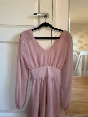 Ljusrosa kortklänning med snörning - Supersöt ljusrosa klänning med v-ringning, långa ärmar och smock vid axlarna. Klänningen har snörning i ryggen och volangdetalj nertill. Perfekt för vår och sommar, tillverkad i ett lätt och luftigt material.