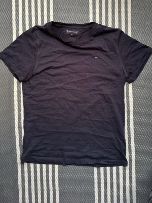 Svart t-shirt från Tommy Hilfiger - En svart t-shirt från Tommy Hilfiger med klassisk passform och liten broderad logga på bröstet. Tillverkad i mjuk bomull och har rund halsringning samt korta ärmar. Perfekt för en stilren och enkel look.
