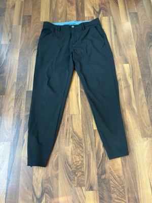 Svarta slim-fit byxor från Macade (golfbyxor) - Svarta byxor från Macade i slim-fit modell. Byxorna har en stilren design med knapp och dragkedja framtill samt diskreta fickor. Perfekta för dig som gillar en modern och clean look. Materialet känns mjukt och följsamt.