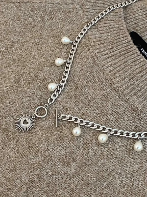 Silverfärgat halsband med pärlor - Unikt halsband i silverfärgad metall med droppformade vita pärlor och en solformad berlock. Kedjan har en chunky design och stängs med ett T-lås. Perfekt för dig som vill ha något som sticker ut och ger en cool touch till din outfit.