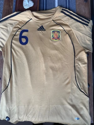 Gul Spanien matchtröja Adidas - Säljer en gul Spanien matchtröja från Adidas med nummer 6 på bröstet och klubbmärke. Tröjan har svarta detaljer, tre ränder på axlarna och UEFA RESPECT-märke på ärmen. Perfekt för fotbollsfans som vill sticka ut på planen.