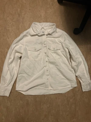 Vit skjorta med tryckknappar - Klassisk vit skjorta i bomull med lång ärm och tryckknappar framtill. Två bröstfickor med lock och knappdetaljer ger en avslappnad men stilren look. Perfekt att styla lager på lager eller bära som den är.