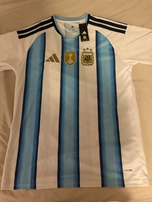 Argentina matchtröja  - Säljer en helt ny officiell Argentina matchtröja från Adidas med klassiska blå och vita ränder. Tröjan har guldiga FIFA- och AFA-emblem på bröstet, samt Adidas-logga. Tillverkad i lätt och ventilerande material. Säljer för att den var för liten.