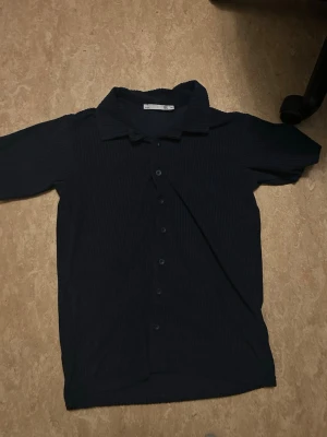 Mörkblå ribbad skjorta med knappar - Säljer en mörkblå skjorta med ribbad struktur och korta ärmar. Skjortan har klassisk krage och knäppning framtill. Perfekt för dig som gillar en enkel men ändå stilren look.