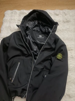 Stone Island jacka - Snygg svart vindjacka från Stone Island med huva och dragkedja. Jackan har två framfickor med dragkedja och den klassiska Stone Island-loggan på ärmen. Materialet är lätt och glansigt, perfekt för blåsiga dagar.