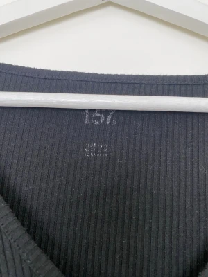 Svart ribbad långärmad topp - En svart ribbad topp med v-ringning och långa ärmar. Toppen har en enkel och stilren design som passar till många olika outfits. Materialet är stretchigt och bekvämt, perfekt för lager på lager eller att bära som den är.