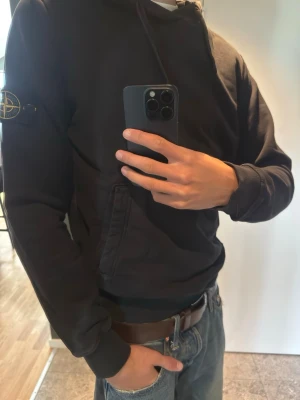 Svart hoodie från Stone Island - Säljer en svart hoodie från Stone Island med klassisk patch på vänster ärm. Hoodien har huva med dragsko, magficka och ribbade muddar. Tillverkad i mjukt bomullsmaterial som är skönt att bära. Perfekt för en avslappnad och stilren look.