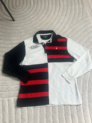 Randig långärmad rugbytröja från Ralph lauren  - Långärmad rugbytröja i 100% bomull med breda svarta och röda ränder, vita partier och klassisk krage. Broderad logga på bröstet och tryck med '9th Division Dist.-14 Champs MCMLXVII. Tröjan är väldigt unik och vintage. perfekt för dig som vill sticka ut och ha en old money, preppy eller vintage-look. Tröjan är XS och passar dig som är emellan 165-170 cm