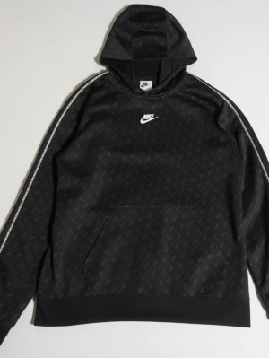 Svart Nike hoodie med mönster - Svart hoodie från Nike med diskret mönster över hela tyget och vit Nike-logga på bröstet. Huva och magficka framtill, samt vita detaljer längs ärmarna. Perfekt för en sportig och avslappnad stil.