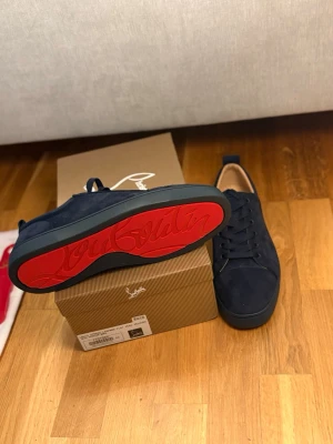 Mörkblå sneakers från Christian Louboutin - Snygga mörkblå sneakers i mocka från Christian Louboutin med den ikoniska röda sulan och diskret logga undertill. Skorna har klassisk låg profil, svarta skosnören och en stilren design som passar till det mesta. Levereras med originalkartong och tillbehör.