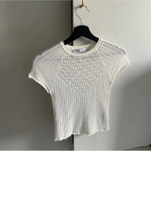 Vit ribbad topp med spetsdetaljer - Zara - Säljer en vit ribbad topp med korta ärmar och fina spetsdetaljer upptill. Toppen är figurnära och har ett snyggt mönster över bröstet som ger en unik look. Perfekt att styla med jeans eller kjol för en trendig outfit.